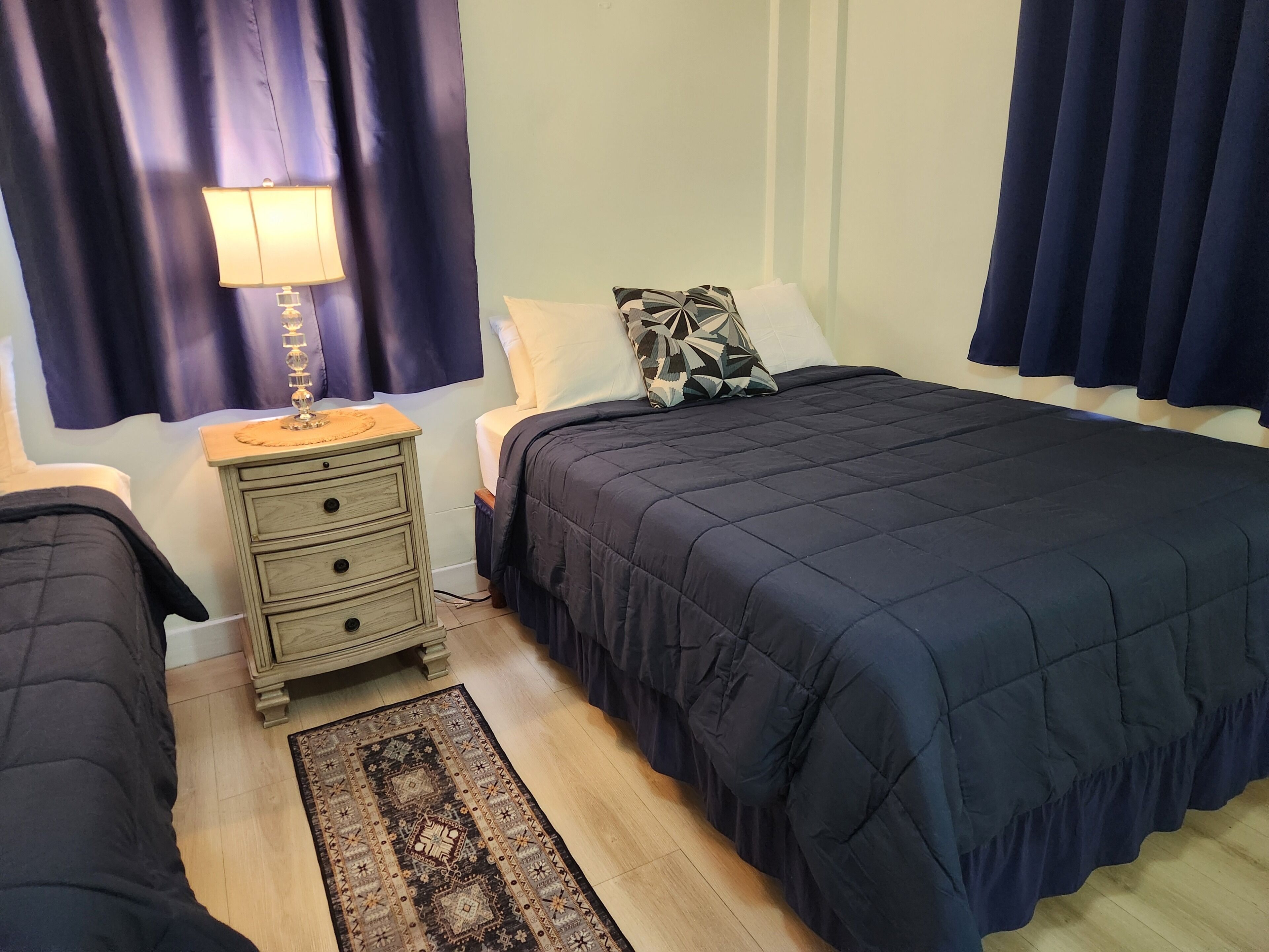 Un dormitorio con cama, mesitas de noche, lámpara, alfombra y cortinas.
