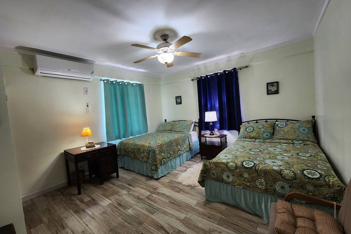 Un dormitorio con dos camas, un ventilador de techo, una mesita de noche con lámpara y suelo de madera.