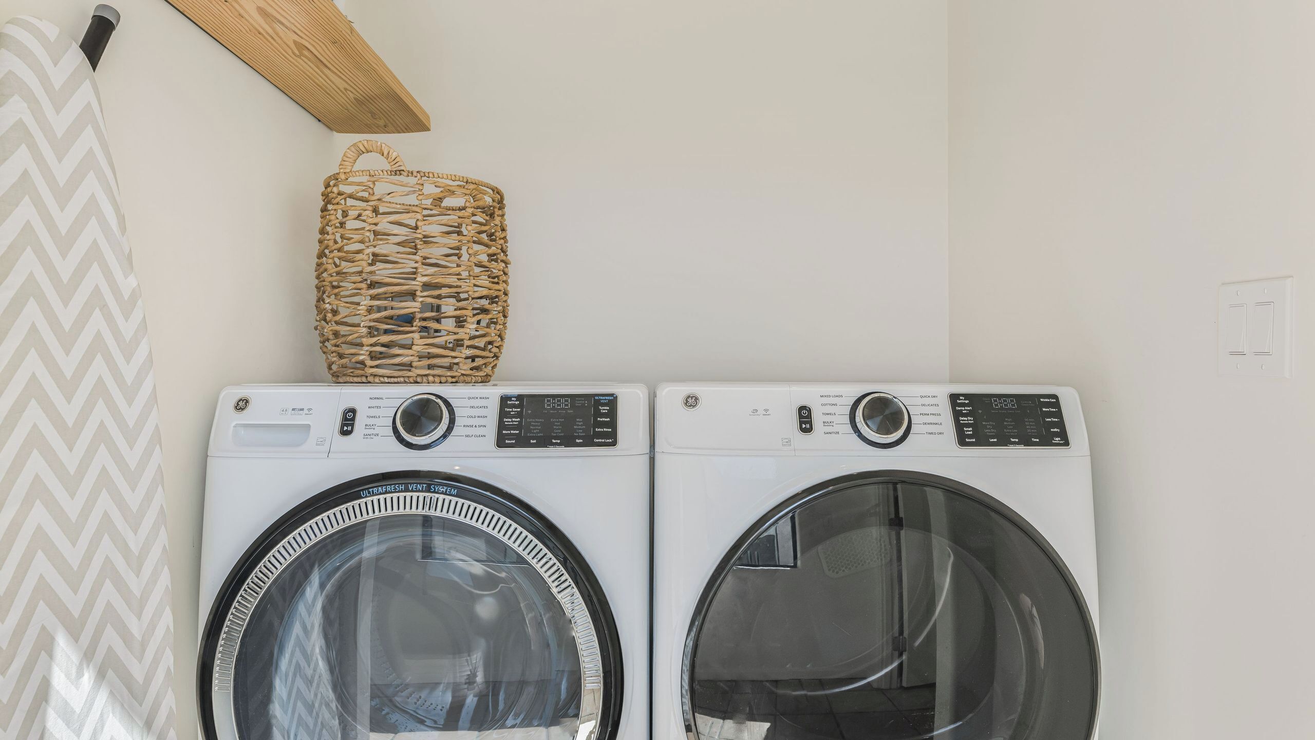 Two front-loading washing machines with control panels, a wooden shelf with a woven basket, and a chevron-patterned towel on the left.