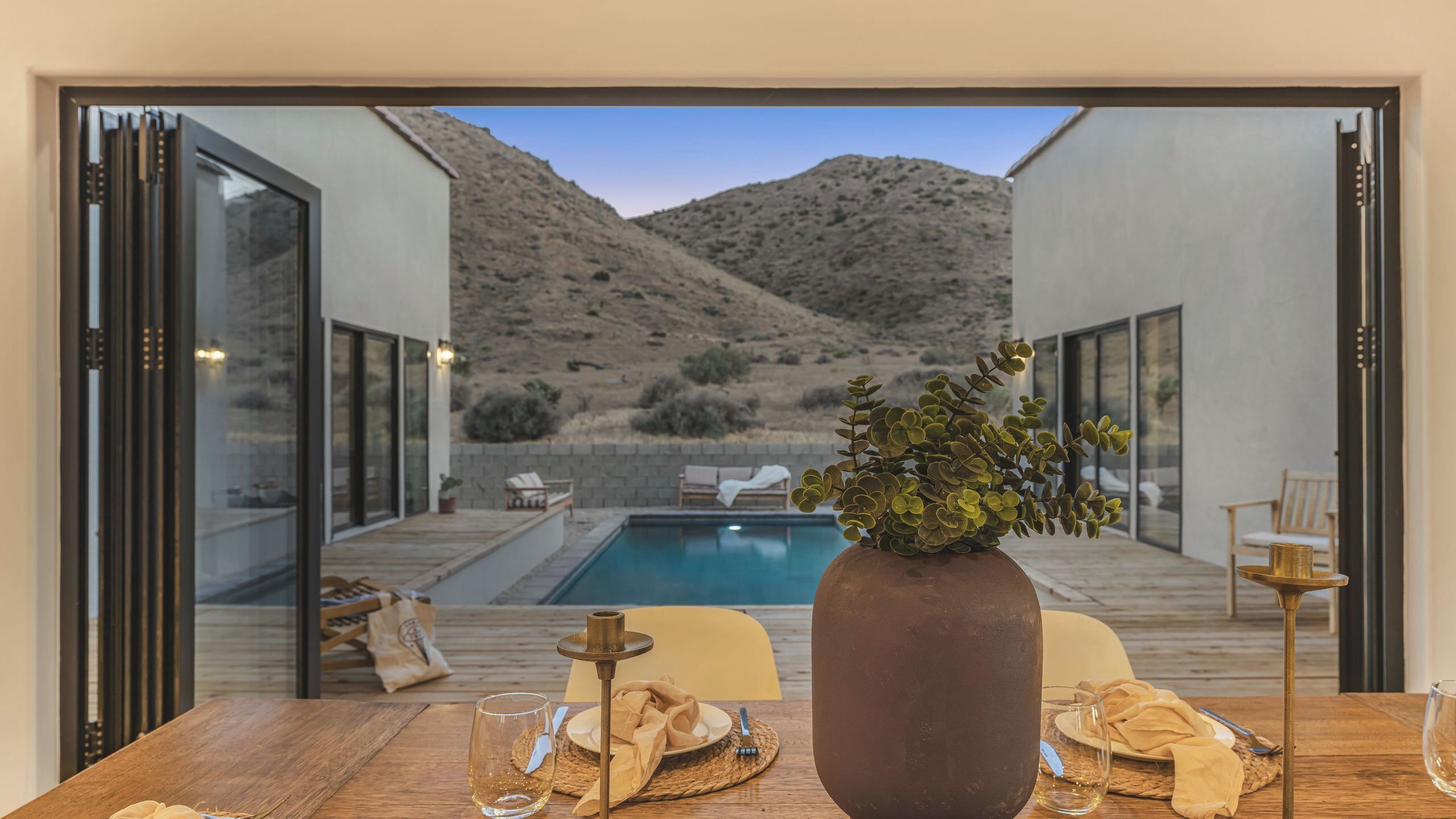 A dining table with a vase of greenery, a glass of wine, and a candle, overlooking a pool and outdoor seating area with a mountainous backdrop.