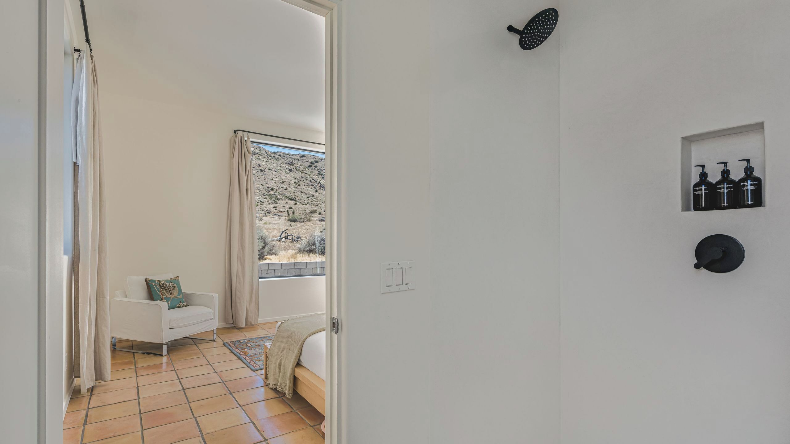 A room with a bed, a window with a view of a hilly landscape, a white wall with a black object and three bottles, and a tiled floor.
