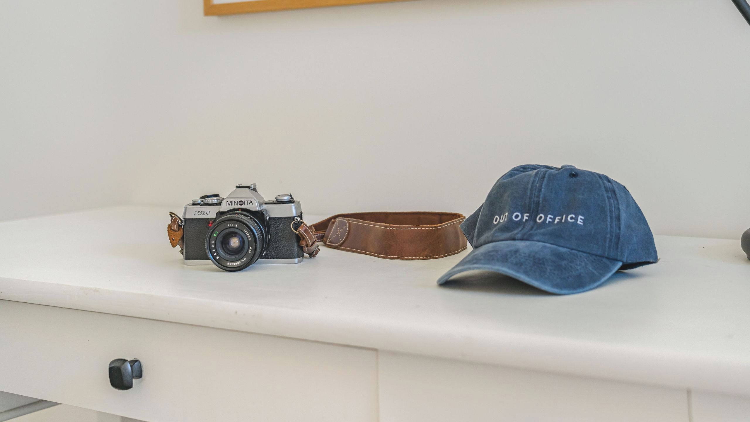 Een oude Minolta X-G1 camera, een lederen band en een blauwe dop met de tekst 'OUT OF OFFICE' op een witte ondergrond.