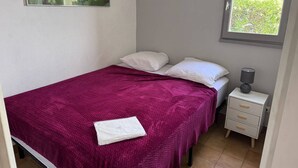 Un dormitorio con cama, almohadas, mesita de noche, lámpara y ventana.