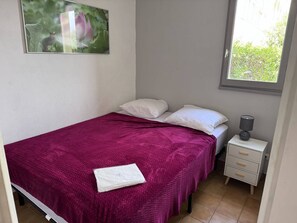 Ein Schlafzimmer mit Bett, Kissen, einem Nachttisch, einer Lampe und einem Fenster.