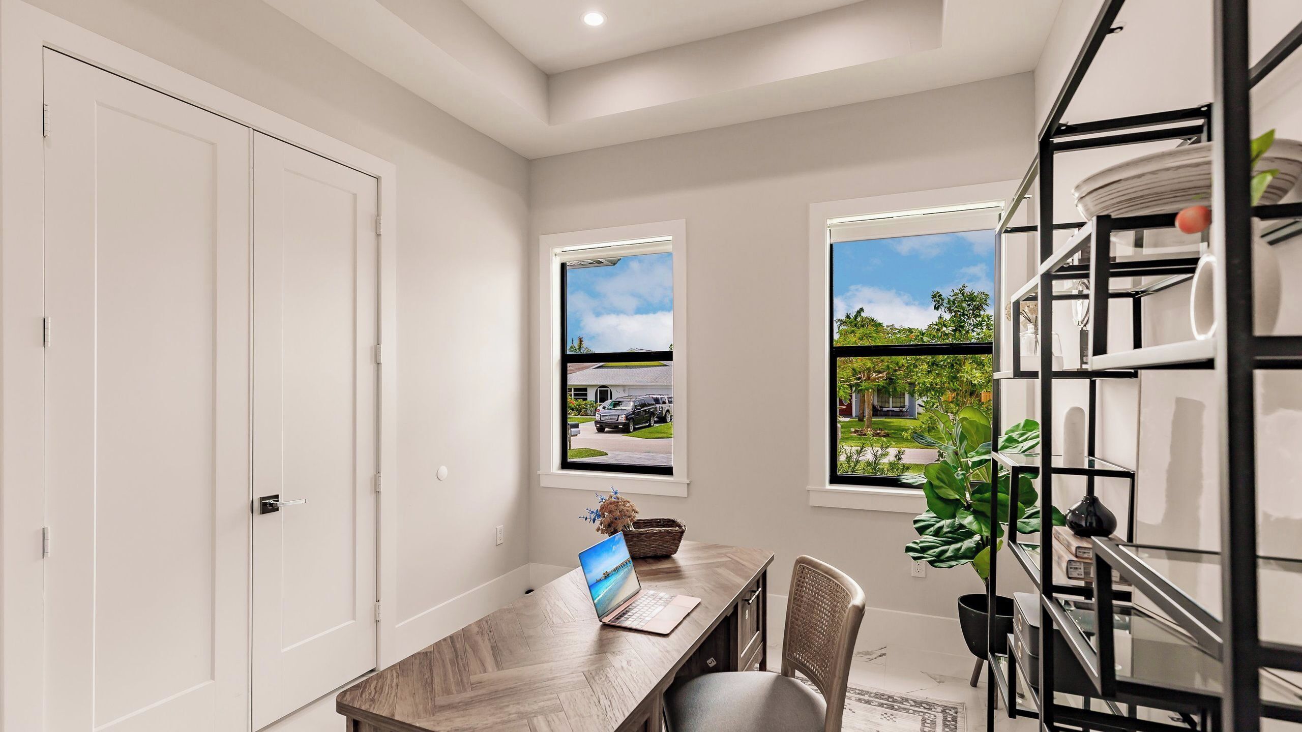 A room with a desk, chair, laptop, two windows with views, and a shelving unit.
