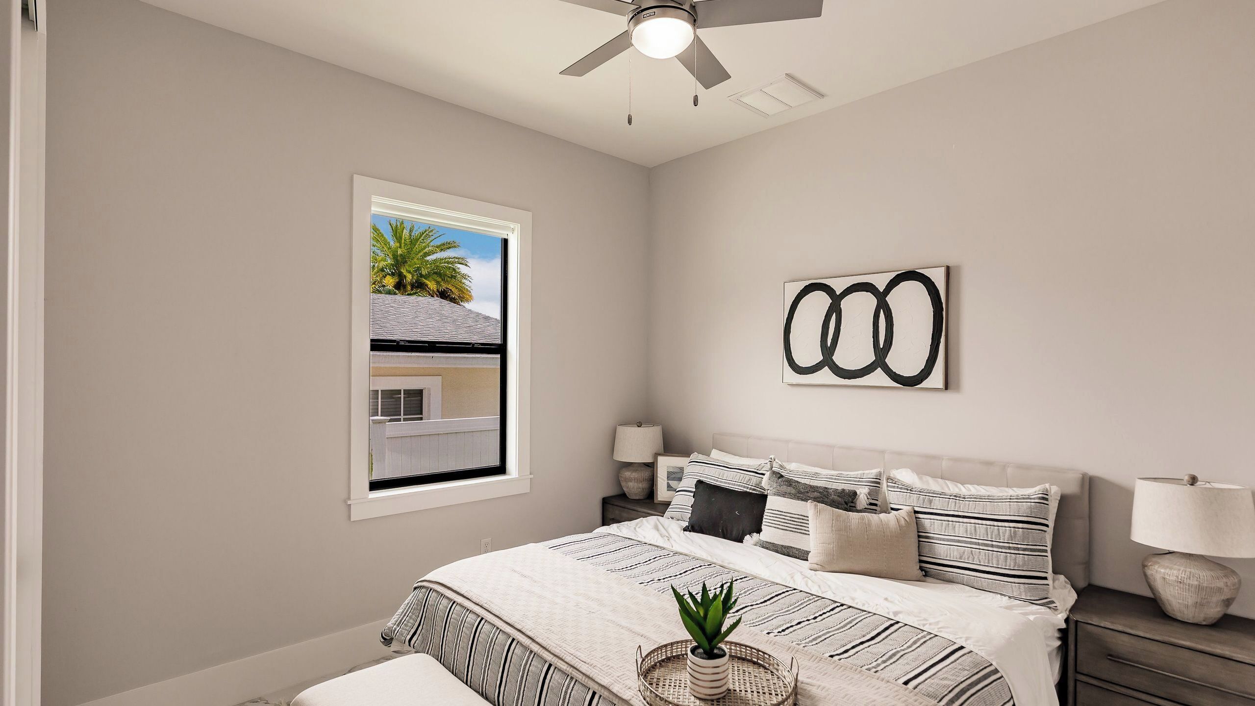 A bedroom with a bed, bedside tables, a ceiling fan, a window with a view, and a wall art piece.