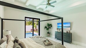 A modern bedroom with a four-poster bed, a TV, a dresser, and a view of a swimming pool. - Coastal Sol by Avantstay - Private Pool, Screened Lanai & Outdoor Kitchen Near Vanderbilt Beach (Naples)