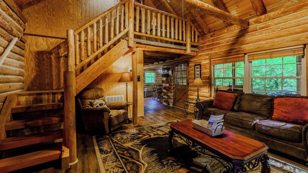 L’intérieur d’une cabane en rondins, avec un escalier en bois, un salon comprenant un canapé et une table basse, et de grandes fenêtres.