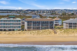 A beachfront hotel with multiple balconies, a sandy beach, and a view of the water and distant town. - Above the Tide | Oceanfront | Community Pool (Nags Head)