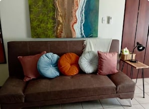 Un sofá marrón con cuatro cojines de varios colores, un cuadro de un paisaje natural y una mesita con una lámpara y una cesta.