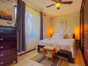 A bedroom with a bed, bedside lamps, a ceiling fan, a dresser, and a wall with a dreamcatcher. - Welcome to CnH Costa Verde: Charming 1-bedroom condo with AC in sunny Coco (Coco)