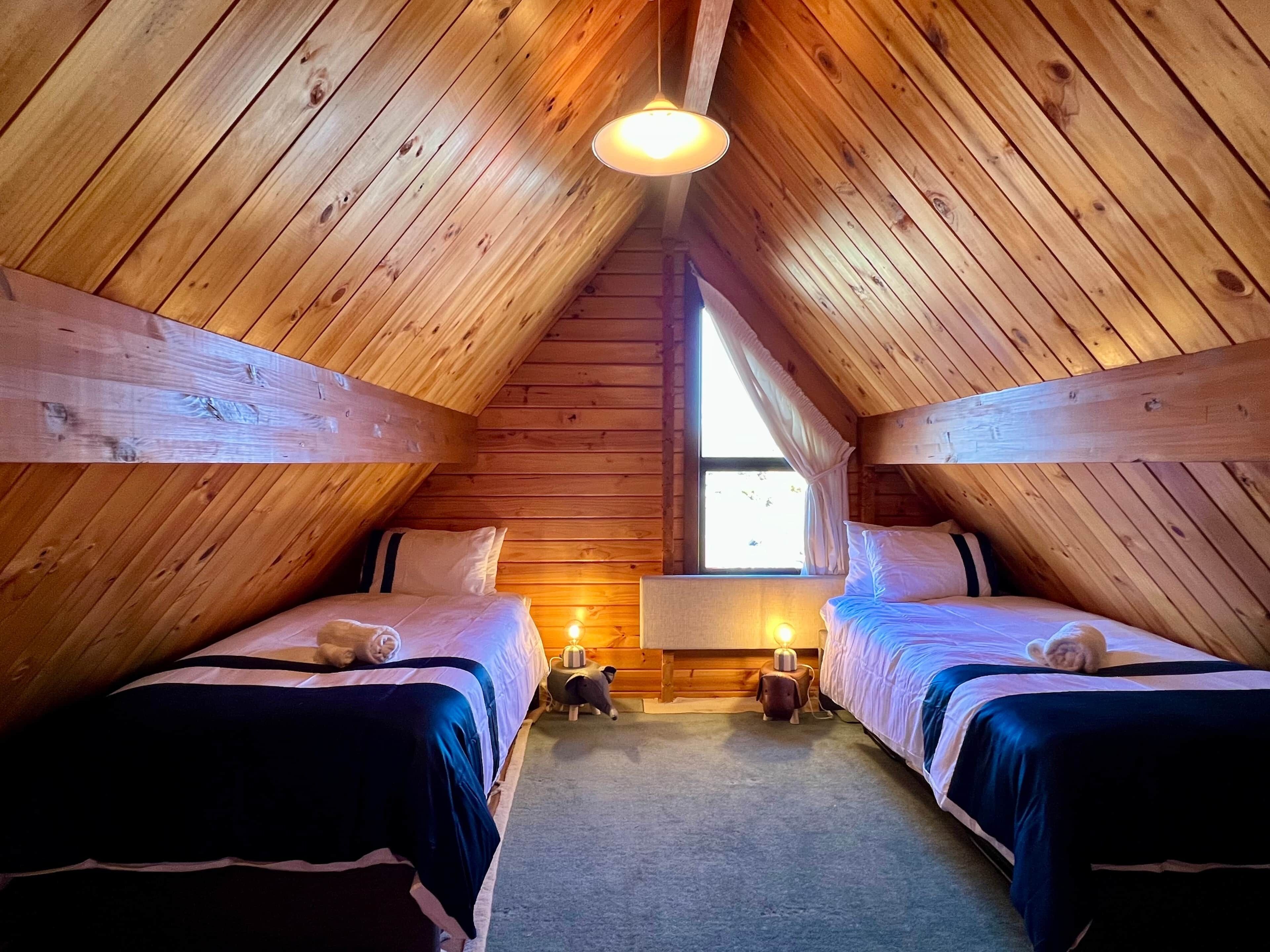 A cozy attic bedroom with wooden walls, two beds, a small window, and a hanging light fixture.