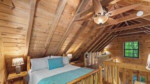Un dormitorio de cabaña de madera con una cama, mesitas de noche, un ventilador de techo y una ventana.