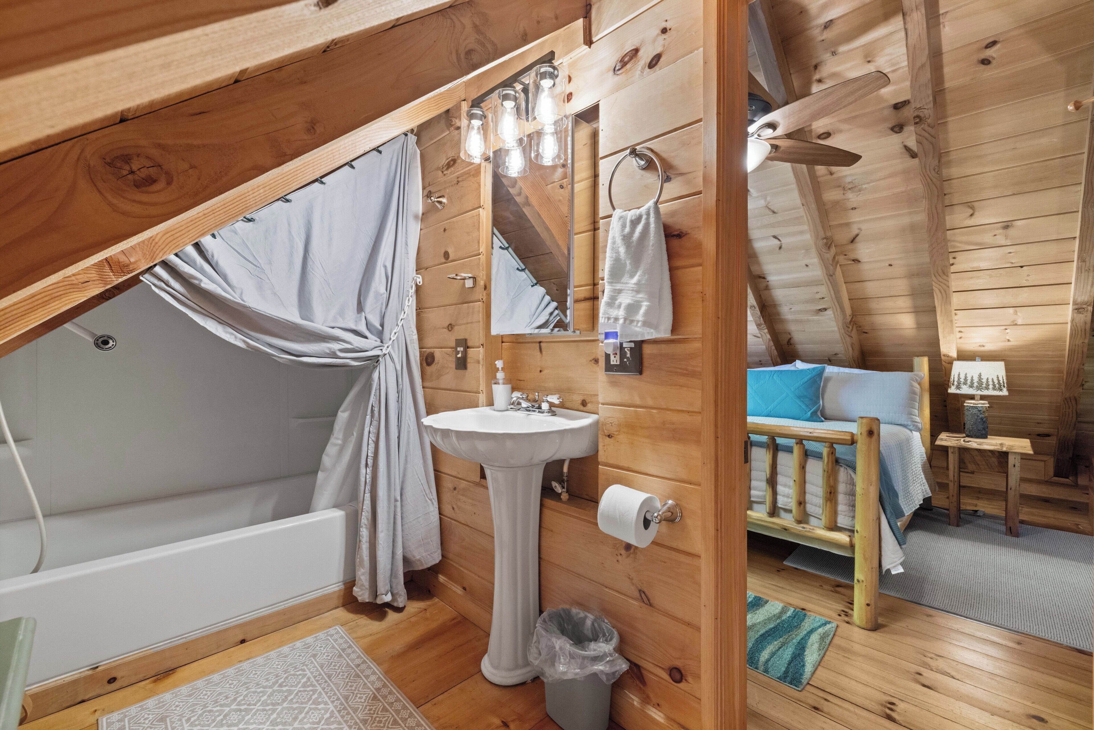 Un dormitori de cabana de fusta amb llit, taula de nit petita, finestra amb cortines i bany amb rentapla, mirall i cortina de bany.
