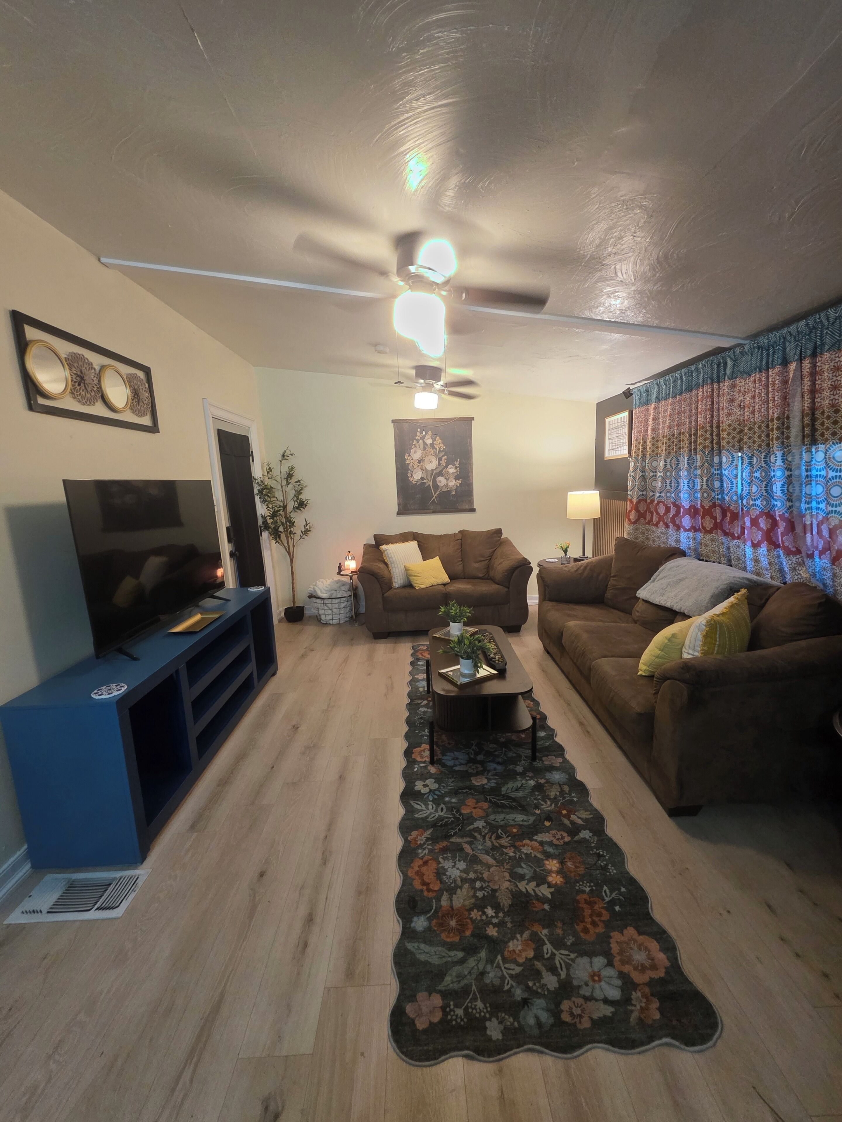 A living room with a flat-screen TV, two couches, a coffee table, a ceiling fan, and a floral rug.