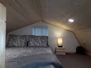 A bedroom with a sloped ceiling, a bed with patterned bedding, a bedside lamp, a small table, and a window with blinds.