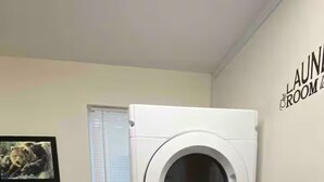 Ein kleiner Waschraum mit gestapelter Waschmaschine und Trockner, Holzschränken, einem kleinen Fenster und einem Wandbild.