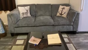 A living room with a grey sofa, two decorative pillows, a black coffee table, and a bear-themed wall art. - The Kodiak Cabin w/ King Bed (Seward)