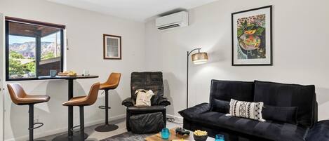 Un salón moderno con un sofá negro, dos sillones marrones, una mesa de centro de madera y vistas a montañas a través de un ventanal grande.