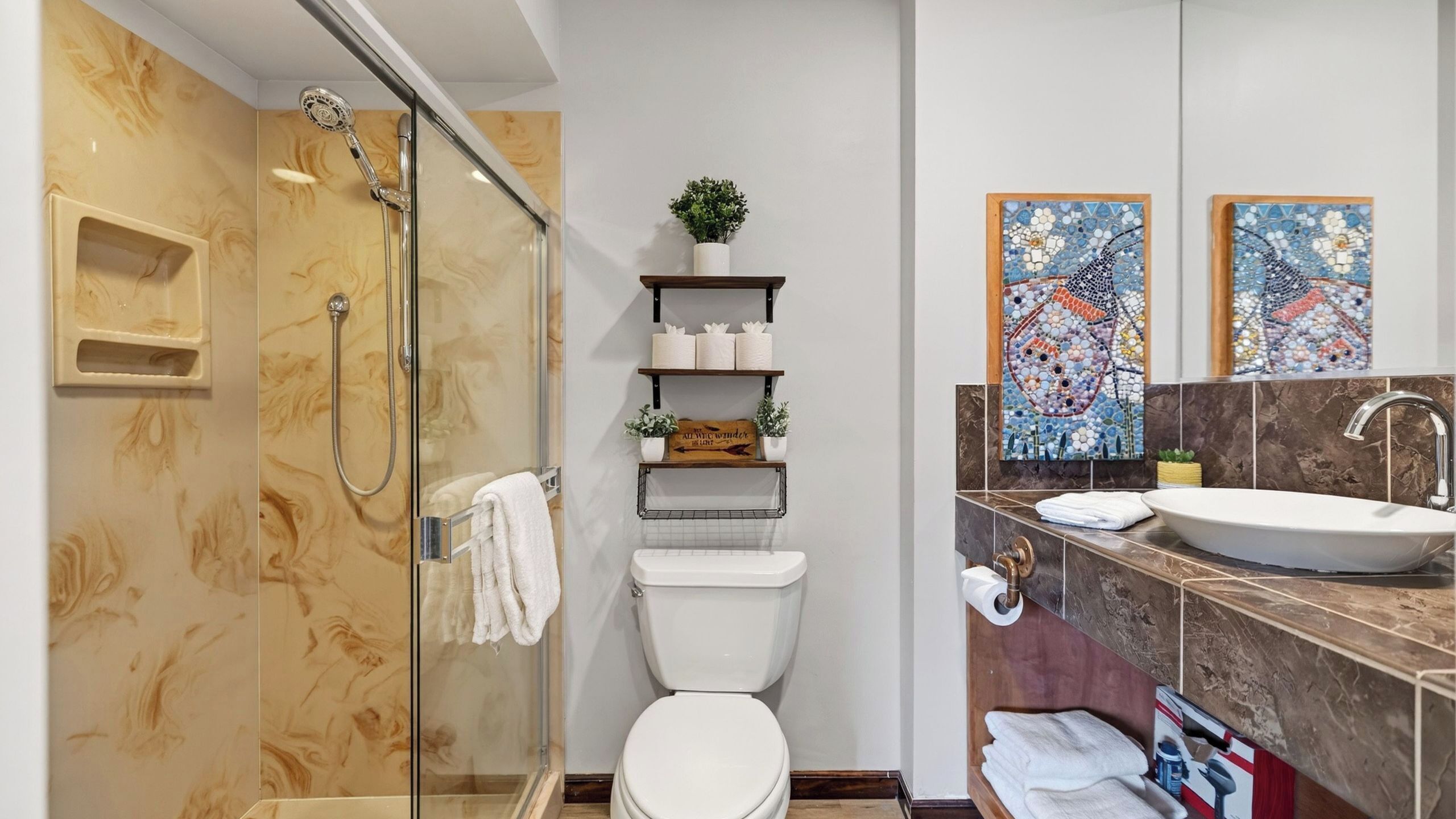 Uma casa de banho moderna com chuveiro em cabine de vidro, sanita, lavatório com bancada de mármore e arte decorativa em mosaico.
