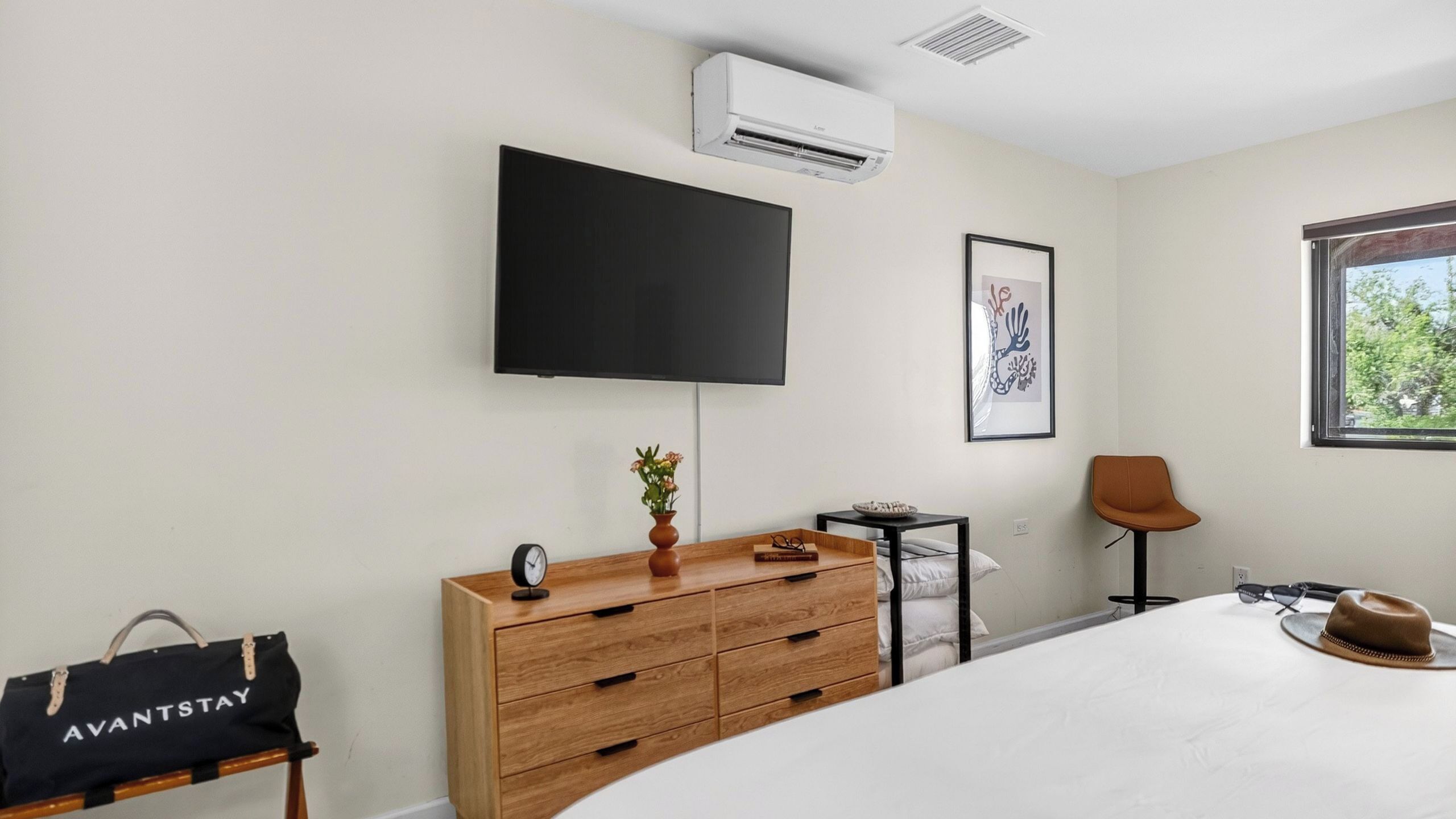 A modern hotel room with a flat-screen TV, a bedside table with a vase and book, a wooden dresser, a chair, and a window with a view.