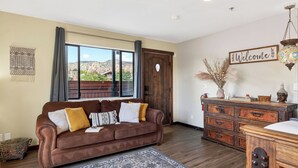 A living room with a brown leather sofa, a wooden dresser, a 'Welcome' sign, and a view of a mountainous landscape through a large window. - Red Rock Duo by Avantstay - Sleeps 8, 2 Sedona Retreats With Mountain Views, Heart of West Sedona (Sedona)
