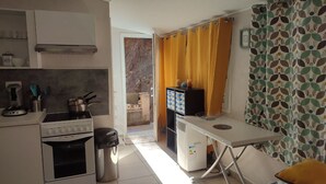Uma cozinha compacta com fogão, forno e máquina de café, uma pequena mesa com uma cadeira e uma cortina estampada.