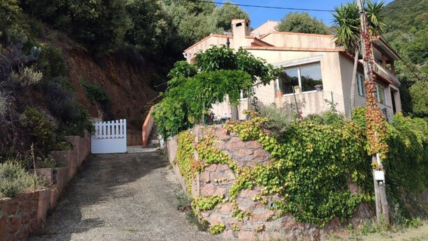 Uma casa com parede de pedra, portão branco e uma colina ao fundo.