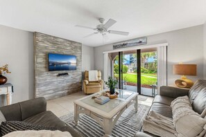 Ein modernes Wohnzimmer mit einem Flachbildfernseher, einem Deckenventilator, einer Couch, einem Couchtisch und Blick auf einen Garten durch Glastüren.