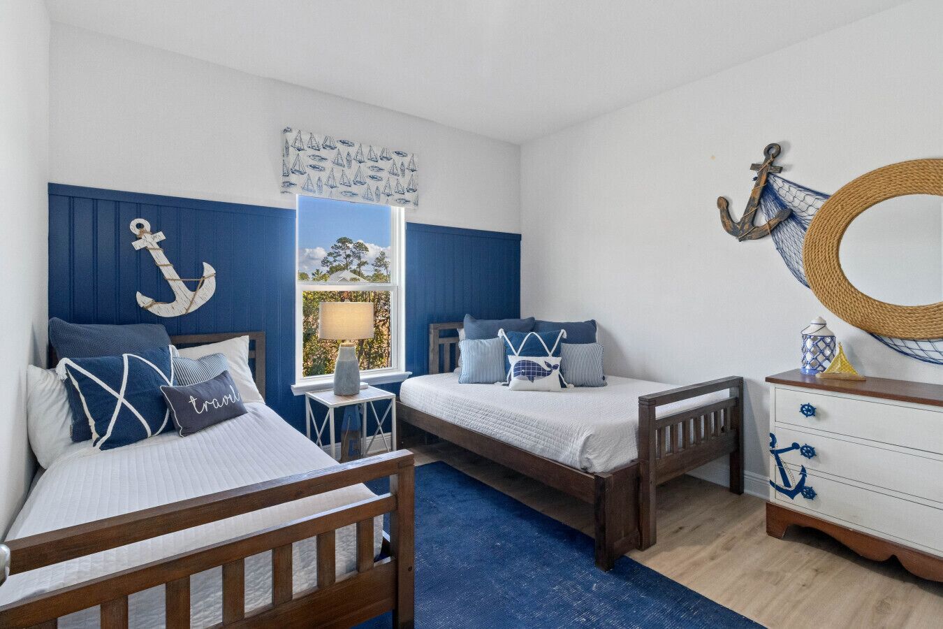 A bedroom with two beds, a window with a view, a dresser with nautical decor, and a wall with an anchor and sailboat artwork.