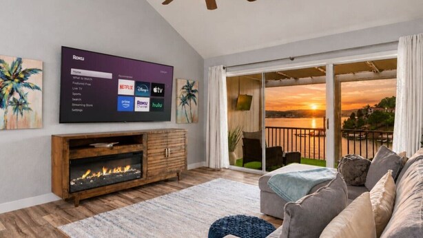 Ein Wohnzimmer mit Flachbildfernseher, der eine Roku-Oberfläche anzeigt, einem Kamin, einer Couch und einem Balkon mit Sonnenuntergangsblick.