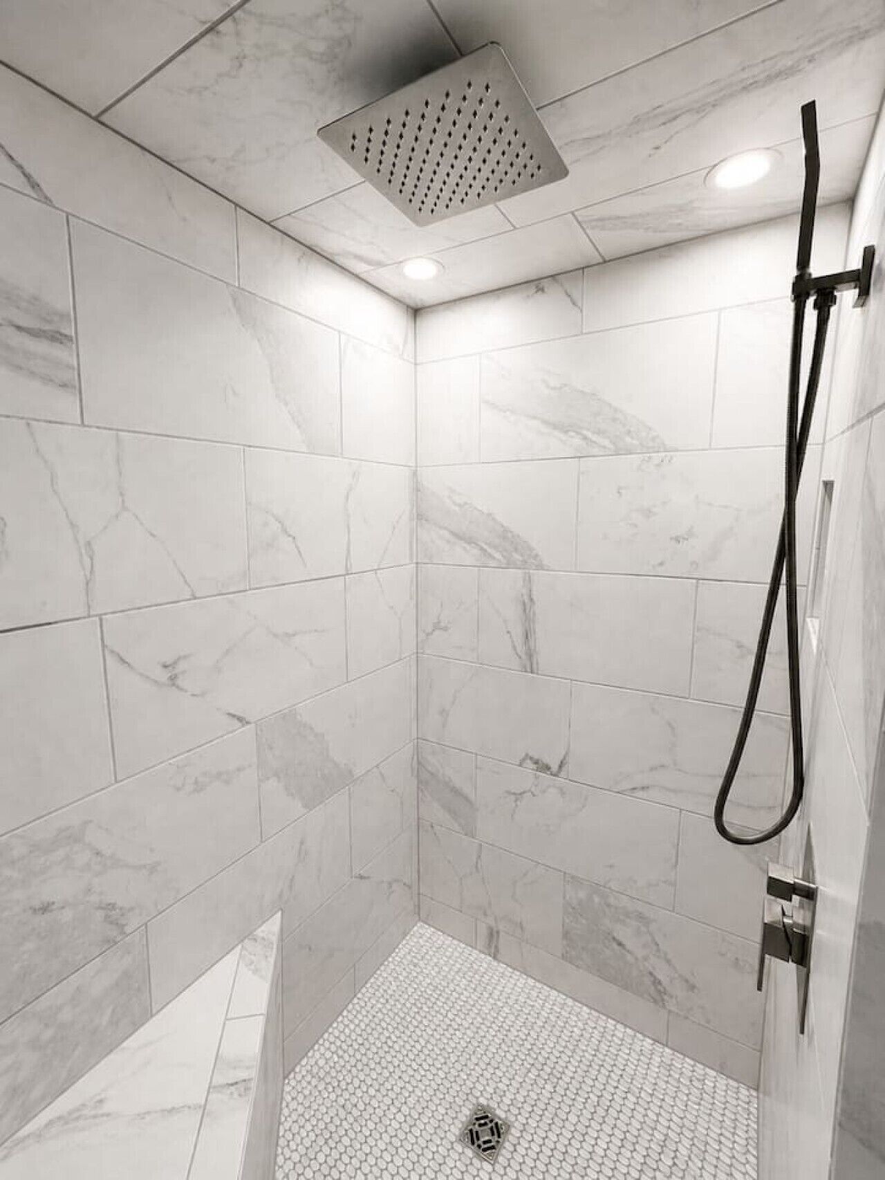 Une douche équipée d’un pommeau de douche flexible, de carreaux de marbre blanc et d’une bouche d’aération au plafond.