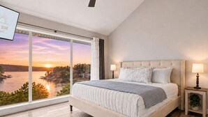Ein Schlafzimmer mit großem Fenster, einem Bett, zwei Nachttischen und Blick auf ein Gewässer bei Sonnenuntergang.