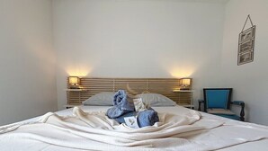 Un lit bien fait, recouvert d’une couette blanche, avec une serviette bleue et un petit objet posé dessus. On aperçoit une tête de lit en bois avec une lampe, une chaise et un cadre photo fixé au mur.