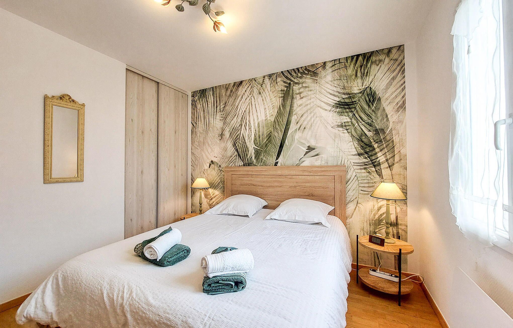 Een keurig opgemaakt bed met handdoeken, een houten hoofdbord, een wand met bladmotief, een nachtkastje met lamp en een spiegel.