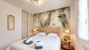 A neatly made bed with towels, a wooden headboard, a wall with a leaf pattern, a bedside table with a lamp, and a mirror on the wall. - Charmante maison proche plage (Saint-Georges-d'Oléron)