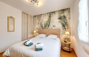 A neatly made bed with towels, a wooden headboard, a wall with a leaf pattern, a bedside table with a lamp, and a mirror on the wall. - Charmante maison proche plage (Saint-Georges-d'Oléron)