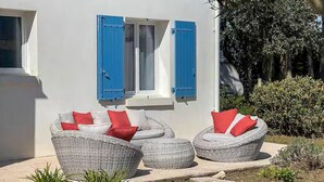 A white building with blue shutters, wicker furniture with red cushions, and a garden area with plants. - Charmante maison proche plage (Saint-Georges-d'Oléron)