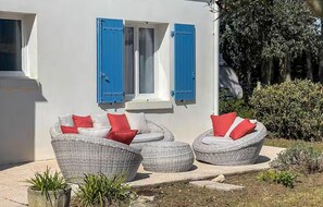A white building with blue shutters, wicker furniture with red cushions, and a garden area with plants. - Charmante maison proche plage (Saint-Georges-d'Oléron)