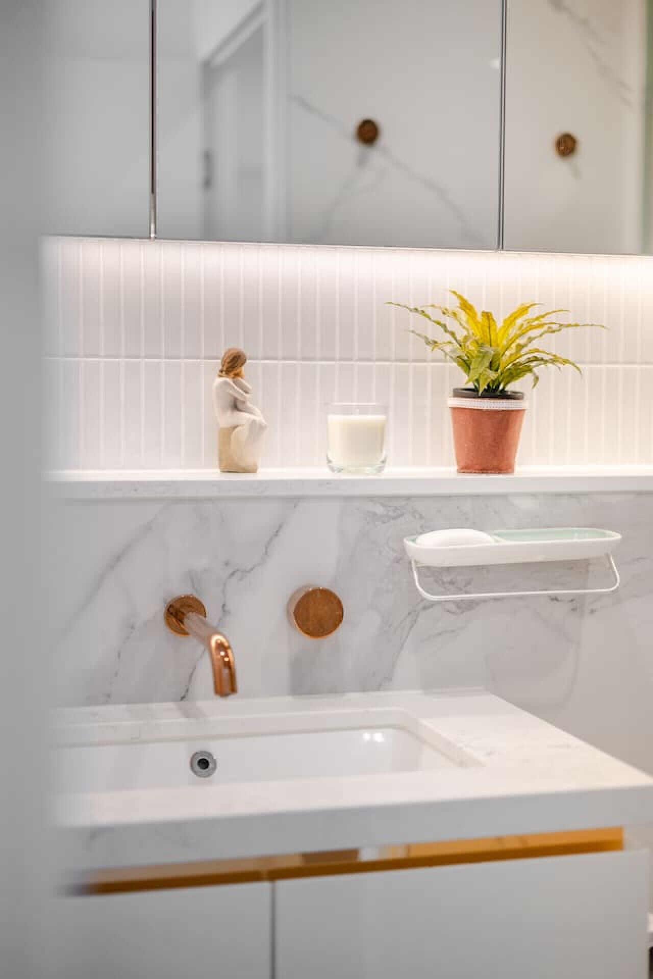 Une salle de bain avec un lavabo blanc, une plante en pot, un porte-bougie en verre et un pare-feu en marbre.