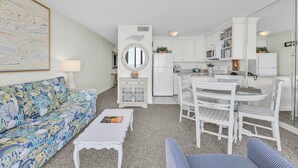 Een compacte woonruimte met een bank, een eettafel met stoelen, een keuken met witte kasten en apparatuur, en een ronde spiegel aan de muur.