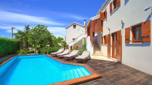 Ein Swimmingpool mit Liegestühlen, ein Holzdeck und ein Gebäude mit weißer Fassade und Holzjalousien.