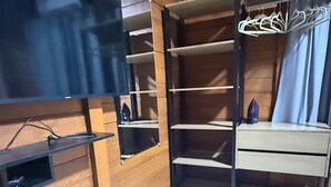 Una habitación con paredes de madera, suelo de baldosas, un televisor en un estante, un armario con espacio para colgar y una mesita.