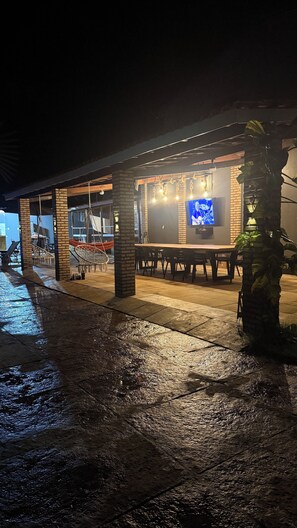 An outdoor seating area with tables and chairs, a TV screen, and hanging lights.