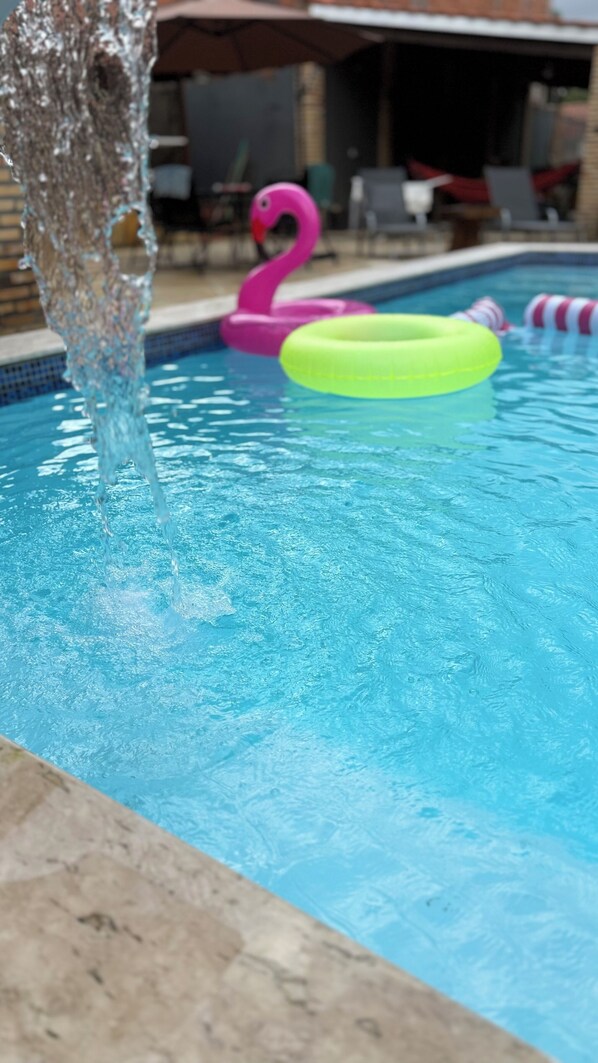 A swimming pool with water flowing into it, a pink flamingo float, and a green circular float.
