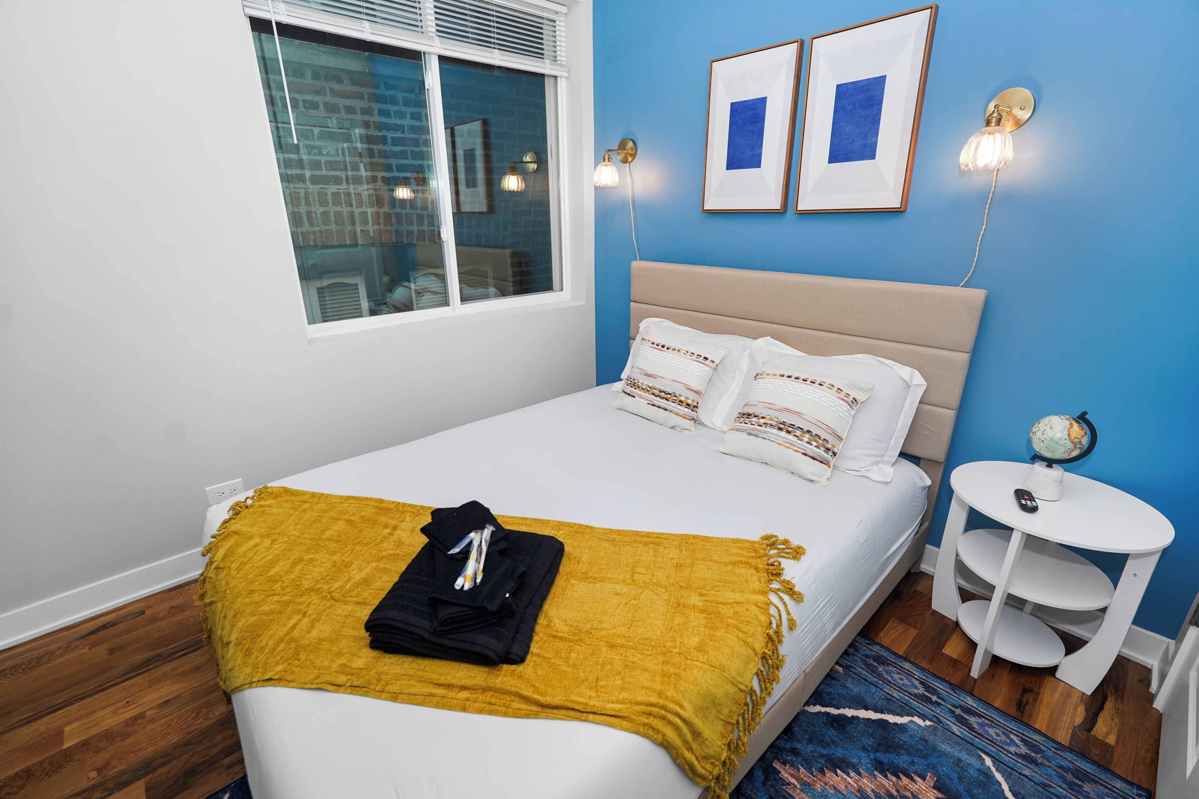 A neatly made bed with a yellow throw blanket, a bedside table with a globe, and a window with blinds.