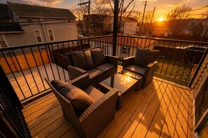Une terrasse dotée de meubles en osier et d’une table en verre, offrant une vue sur un quartier résidentiel au coucher du soleil.