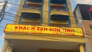 Un edificio de varios pisos con una fachada llamativa de color amarillo y azul, un letrero iluminado grande que dice 'KHÁCH SAN SÔN TINH' y una entrada visible con puerta de vidrio.