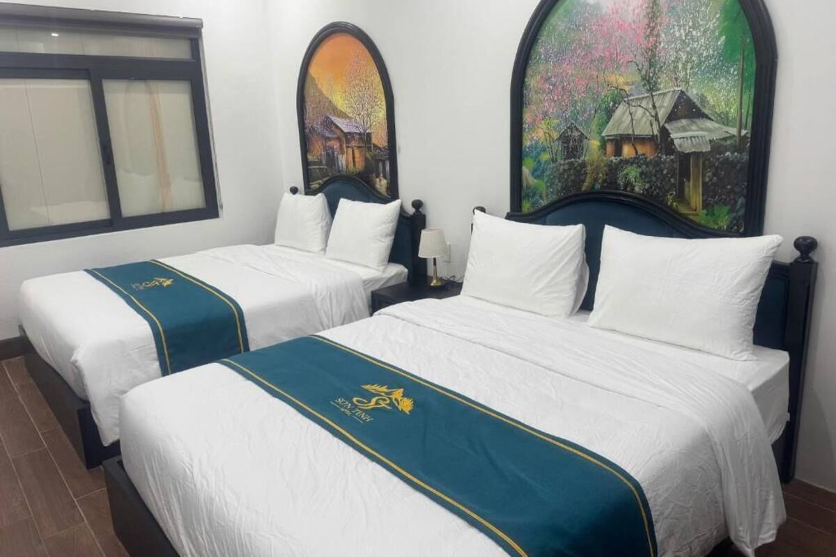 A hotel room with two beds, each with white bedding and teal bedspreads. There are two paintings on the walls, one depicting a village scene and the other a colorful landscape.