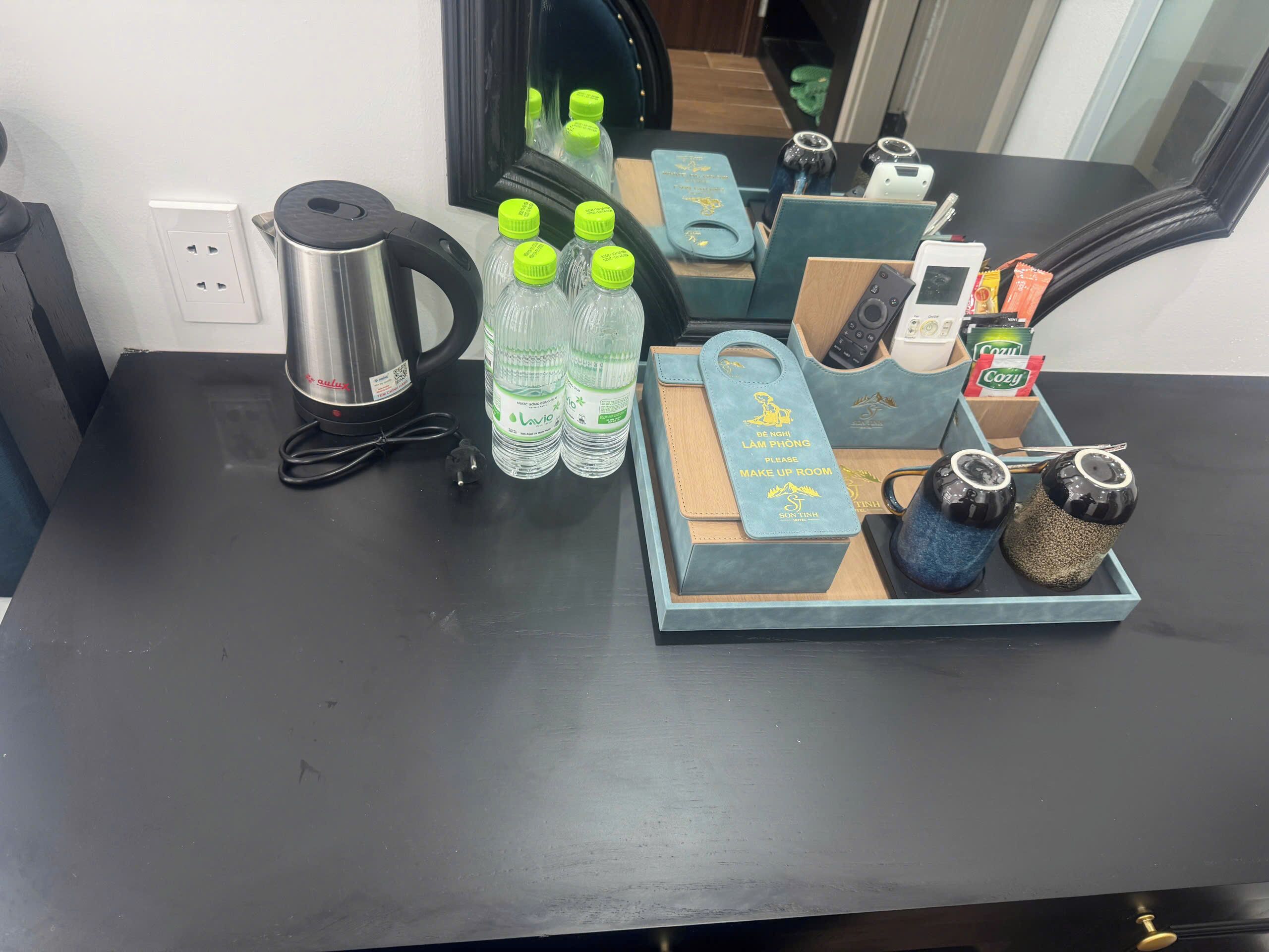 A hotel room desk with a kettle, water bottles, a tray with a towel and salt and pepper shakers, a remote control, and a box of Cozy tea.